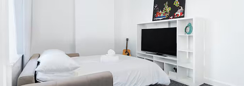 modern bedroom with white walls featuring a bed and a television stand showcasing minimalistic decor and a guitar in the corner cozy atmosphere for relaxation